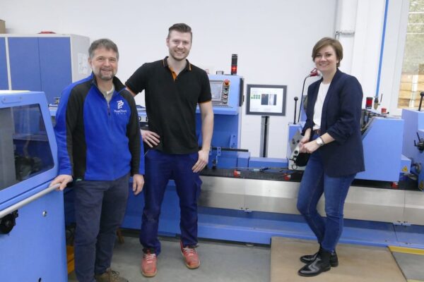 Drei Personen stehen vor einer blauen Weiterverarbeitungsmaschine in der Werkstatt
